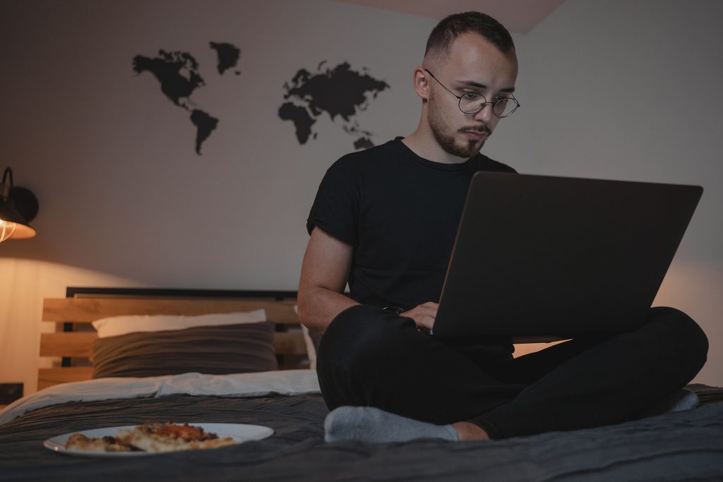 Man in black crew neck t shirt sitting on bed using a laptop, representing UCSTAK's E911 for Remote, Hot-Desk & Field Workers feature.